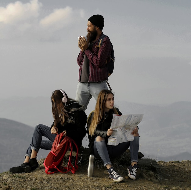 Young friends travelers bearded man hipster and two pretty girls women with map explore route outdoors on mountain top on foggy sky