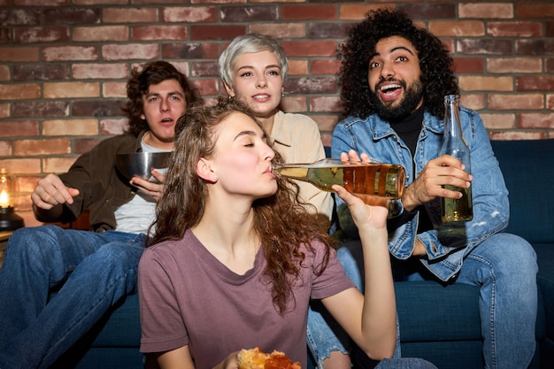 Young friends watching tv, comedy show or movie, eating snacks and drinking beverage, sitting on cozy couch at home, diverse guys and ladies enjoying free time, weekend together