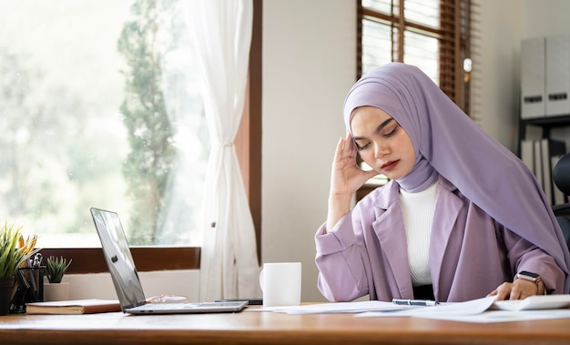 Young frustrated arabic Muslim young woman in hijab working at office desk in front of laptop suffering from chronic daily headaches