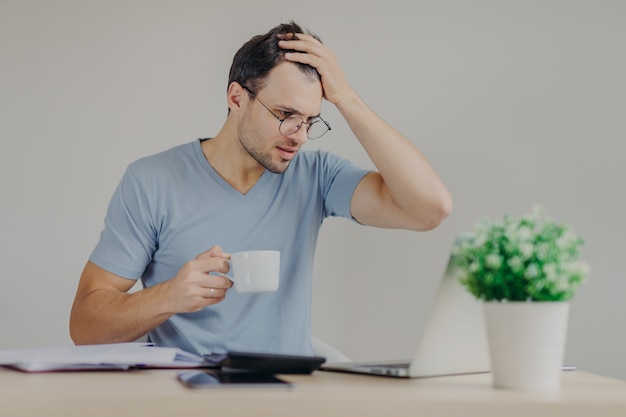 Young frustrated male does paperwork, reads with puzzled expression unpleasant information on laptop computer