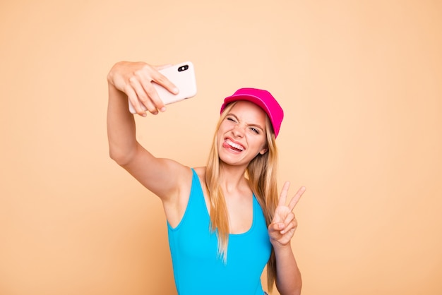 Young funky girl showing v-sign tongue out making self picture with smart phone