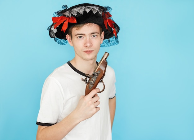 Young funny male teenager in white t-shirt on blue background in pirate hat and gun in hand