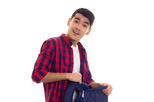 Young funny man with black hair in white Tshirt and red checkered shirt holding blue backpack