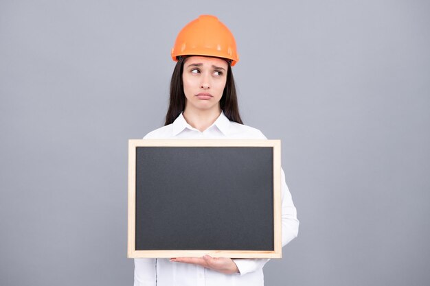 Young funny sad woman construction manager Woman builder isolated portrait with protect helmet and board Gray background empty space copy space