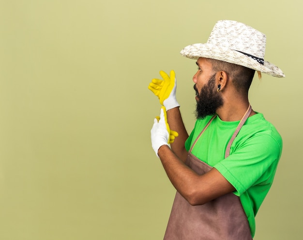 Young gardener afro-american guy wearing gardening hat with gloves points at behind 