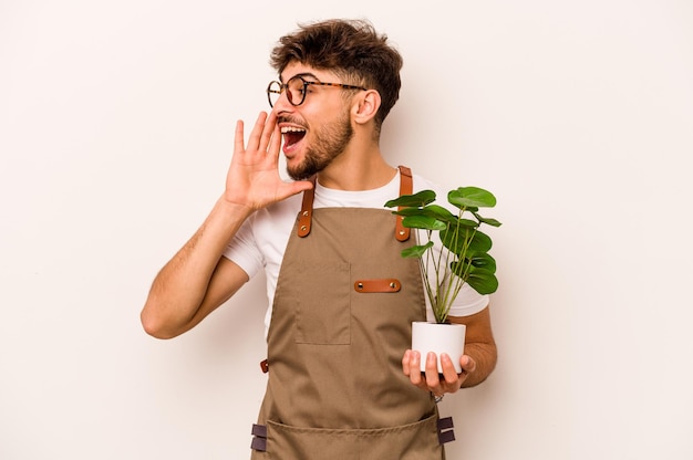 Young gardener hispanic man holding a plant isolated on white background shouting and holding palm near opened mouth