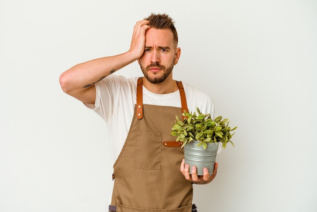 Young gardener tattooed caucasian man holding a plant isolated on white background being shocked, she has remembered important meeting.