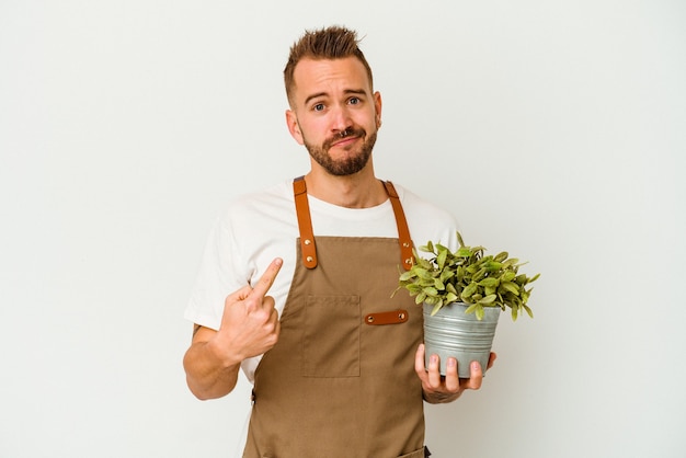 Young gardener tattooed caucasian man holding a plant isolated on white background pointing with finger at you as if inviting come closer.