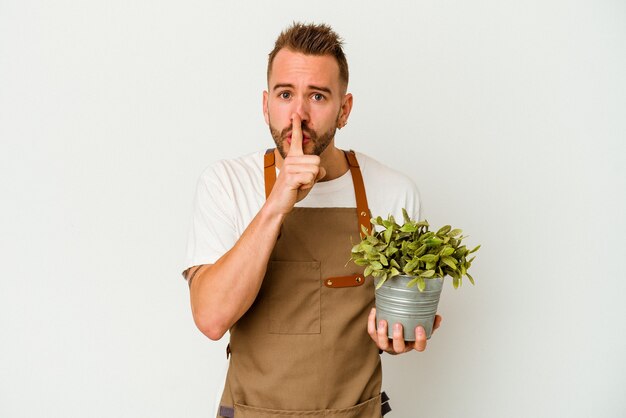 Young gardener tattooed caucasian man holding a plant isolated on white wall keeping a secret or asking for silence.