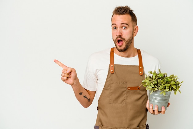 Young gardener tattooed caucasian man holding a plant isolated on white wall pointing to the side