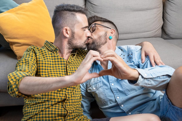 Young gay couple kissing while doing heart symbol with hands at home