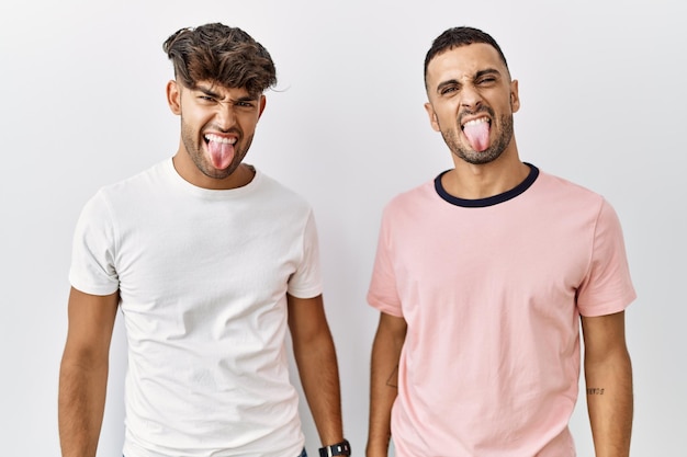 Young gay couple standing over isolated background sticking tongue out happy with funny expression emotion concept