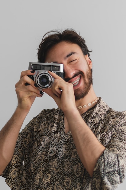 Young gay latin man smiling and with painted nails taking pictures with an antique camera with a white background Vertical photo