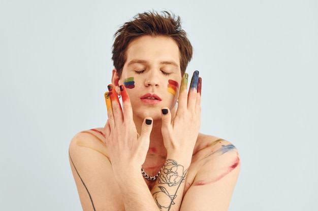 Young gay man is standing in the studio and posing for a camera Shirtless guy with rainbow make up