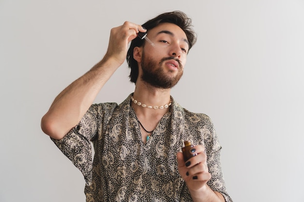 Young gay queer latin man applying oils to his face with an eyedropper on a white background