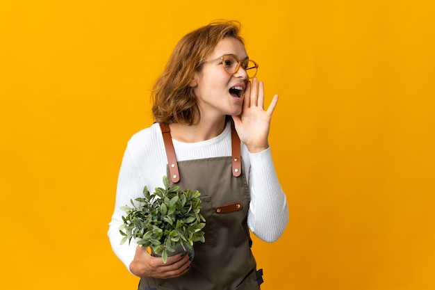 Young Georgian woman holding a plant isolated on yellow wall shouting with mouth wide open to the side