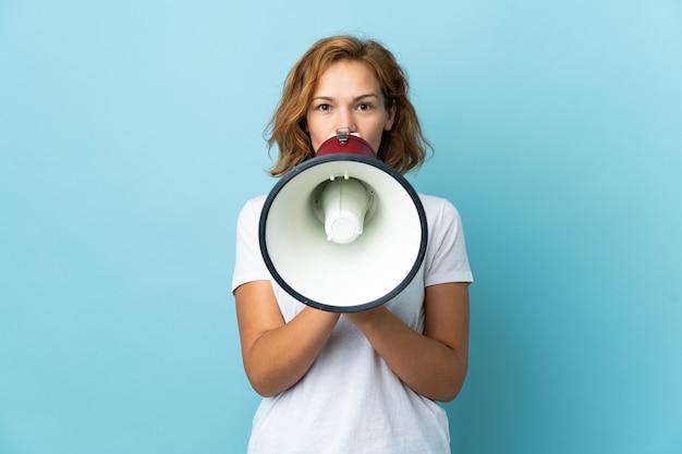 Young Georgian woman isolated on blue wall shouting through a megaphone to announce something