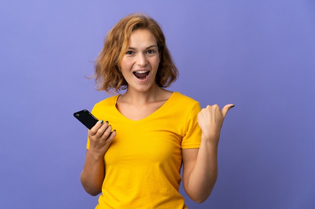 Young Georgian woman isolated on purple wall using mobile phone and pointing to the lateral