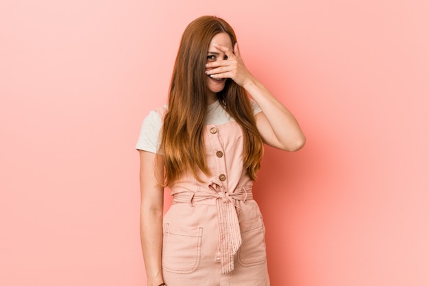 Young ginger woman with freckles blinking through fingers