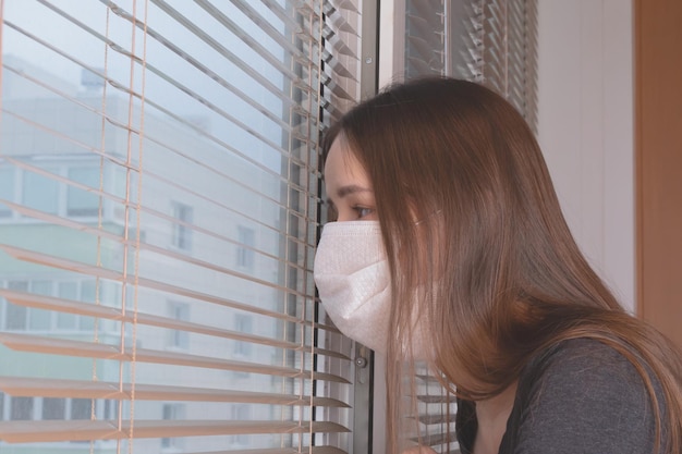 A young girl in an antibacterial mask looks out the window from the house to the street