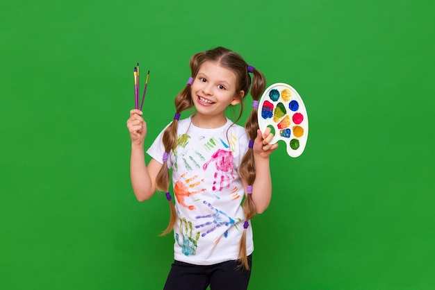 A young girl artist with paints and a brush on a green background in a white Tshirt Children's creativity and drawing with paints