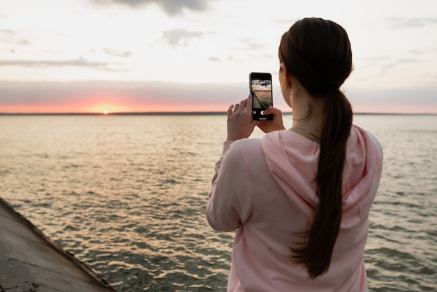 A young girl, an athlete, after a bike ride, came to the sea, photographs the dawn, looks at the sea and rests from doing sports.