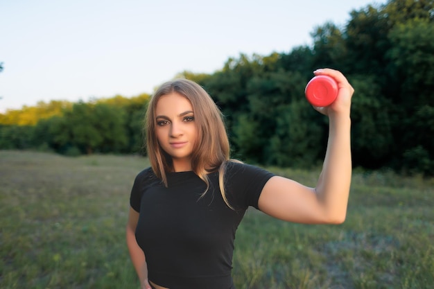 A young girl athlete holds a red bottle of water in her hand showing her muscles on her arm