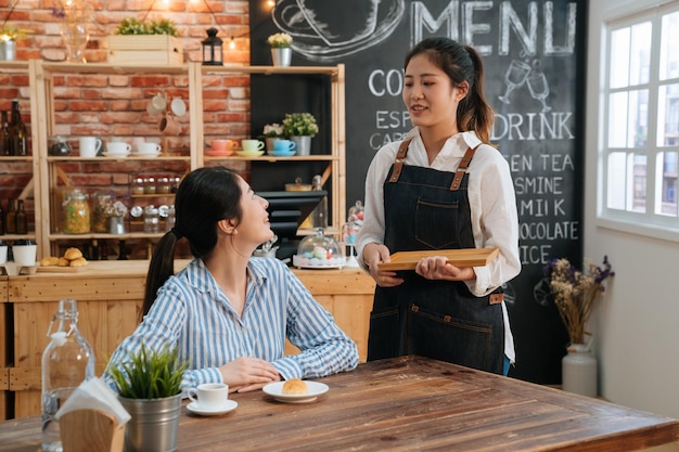 young girl barista in apron chatting with guest at cafe table while sending meal. Woman offer fresh coffee and croissant to customers at modern bar interior. occupation people and service concept