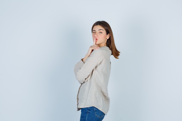 Young girl in beige shirt, jeans showing silence gesture, winking and looking cute , front view.