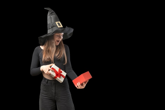 Young girl in black clothes and pointed hat holds an open gift and looks at it in surprise Portrait witch girl isolated on black background