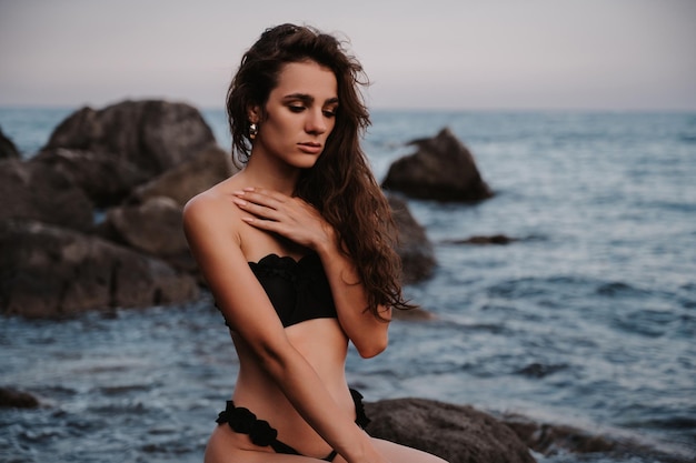 Young girl in a black swimsuit is sitting sexy on rocks on the beach by the sea in summer