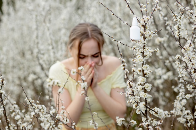 Young girl blowing nose and sneezing in tissue in front of blooming tree. Seasonal allergens affecting people. Beautiful lady has rhinitis.