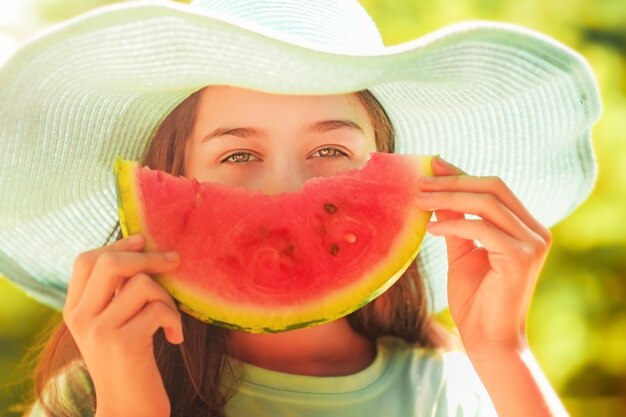 Young girl in a blue hat holding a slice of watermelon on a summer background. Vegetarian