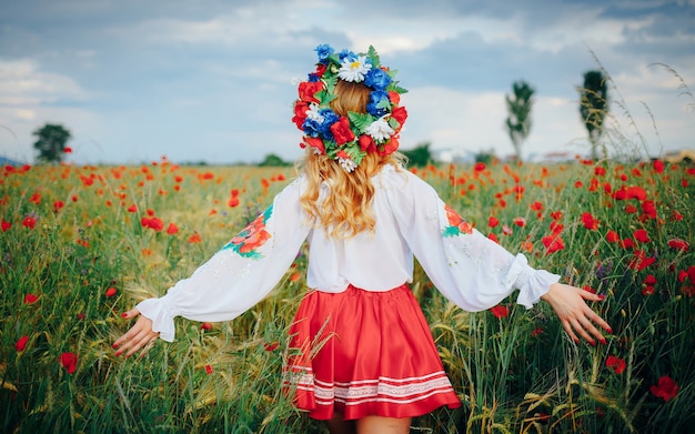 A young girl in bright national clothes and with a wreath of flowers on her head, spreading her arms, walks across a field of red poppies