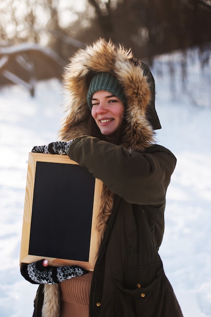 A young girl, brunette, frame in hands, blackboard, copy space, against backdrop of the winter landscape. Snow and frost, the concept of Christmas.