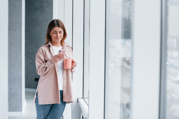 Young girl in casual clothes standing indoors with her phone in hand