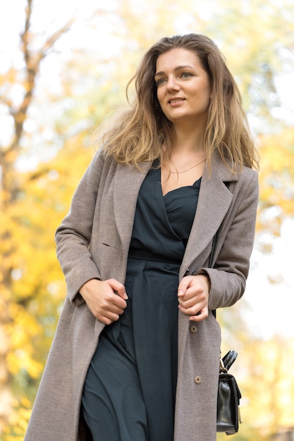 A young girl in a coat poses in a park on a background of golden autumn foliage.