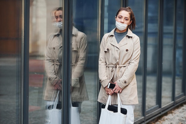 Young girl in coat and white protective mask standing outdoors near building