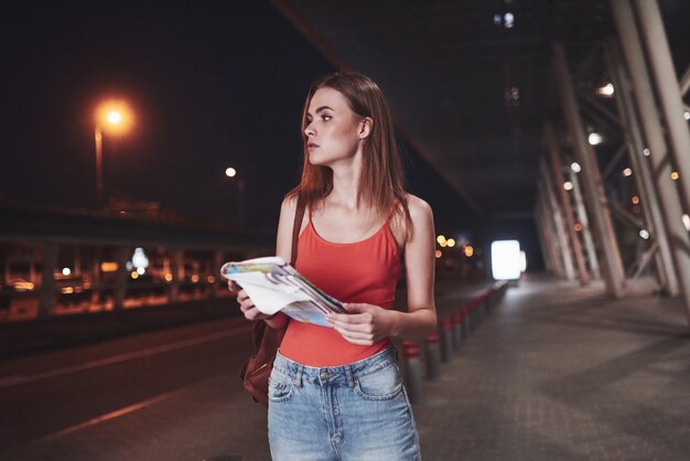 Young girl costs at night near the terminal of the airport or station and reading city map