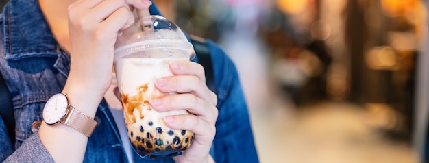 Young girl in denim jacket is drinking brown sugar flavored tapioca pearl bubble milk tea with glass straw in night market of Taiwan, close up, bokeh