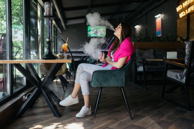 Young girl drinking aperol Aperol spritz cocktail in a glass The girl smokes a hookah Hookah in a restaurant Woman smokes a hookah and sits on a chair