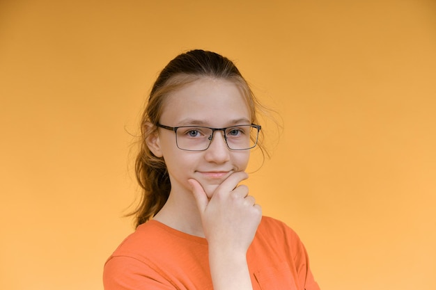 A young girl in glasses holds her face with her hand and looks thoughtfully into the camera Portrait of a girl on a yellow background