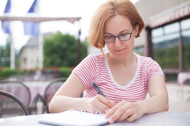 A young girl in glasses sits at a table and makes notes in a notebook, a light warm summer mood