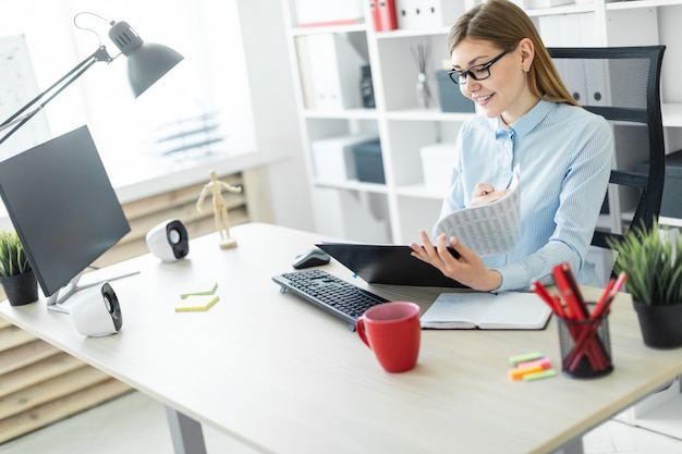 A young girl in glasses sits at a table in the office, holds a pencil in her hand and works with documents.