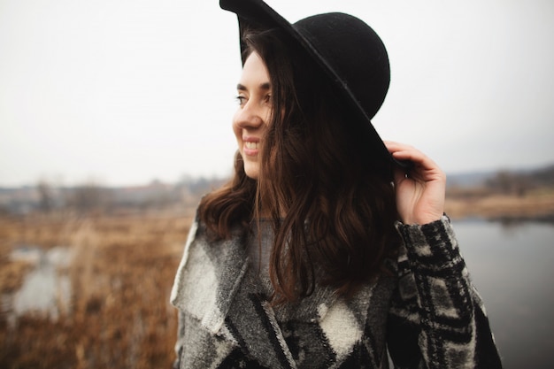 Young girl in gray cardigan and black hat smiles and poses on the shore of a lake