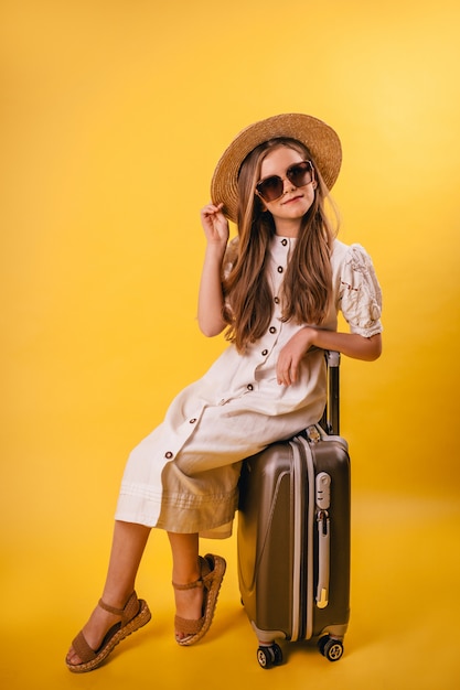 Young girl in a hat sits on a brown suitcase with a yellow background