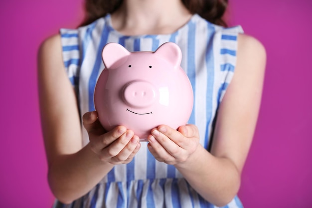 Young girl holding cute piggy bank in hands on pink background Saving for education concept