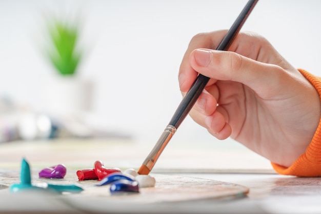 A young girl holds a paint brush in her hand and dunks it in oil paint. 