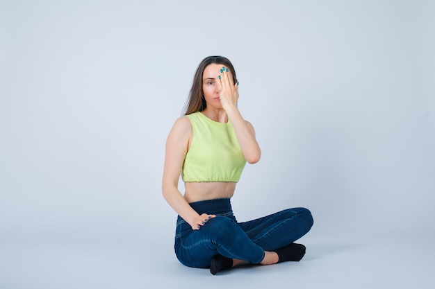 Young girl is covering her half face with hand by sitting on floor on white background