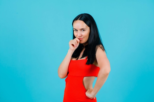 Young girl is looking at camera by holding hand on mouth and putting other hand on waist on blue background